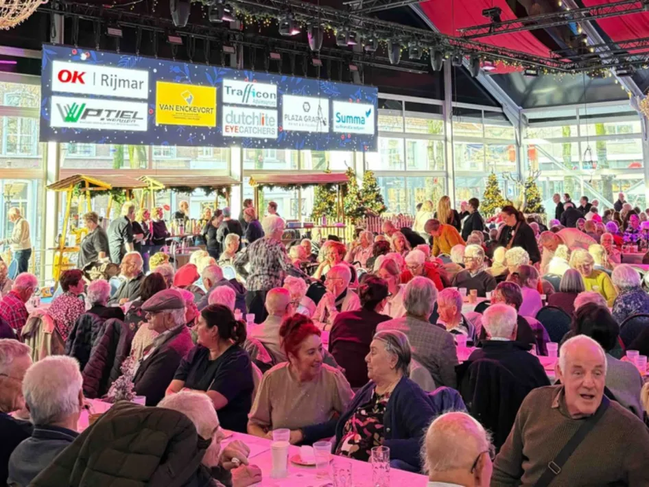 Een drukbezochte bijeenkomst in een sfeervol winterpaviljoen, waar senioren samen aan lange tafels zitten en genieten van eten, drinken en gezelligheid.