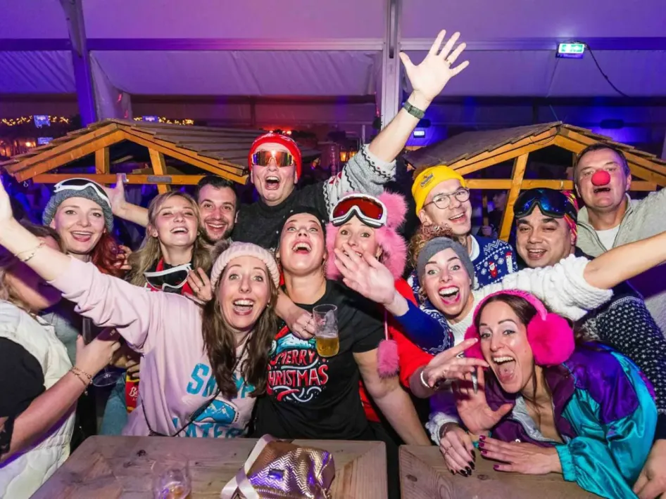Een groep feestgangers in winterse outfits, met skibrillen, mutsen en kleurrijke kleding, poseert tijdens de Après Ski Party in de Winter Fever Arena.
