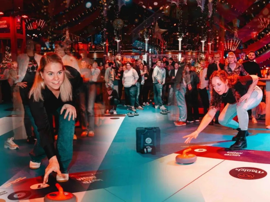 Twee vrouwen spelen fun curling op indoor curlingbanen tijdens de Fun Curling Cup in de Winter Fever Arena, terwijl bezoekers op de achtergrond toekijken.