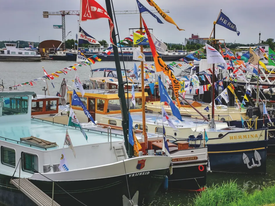 Versierde schepen aangemeerd in de haven van Maasbracht tijdens de Maasbrachter Havendagen, met kleurrijke vlaggen en bezoekers langs het water.