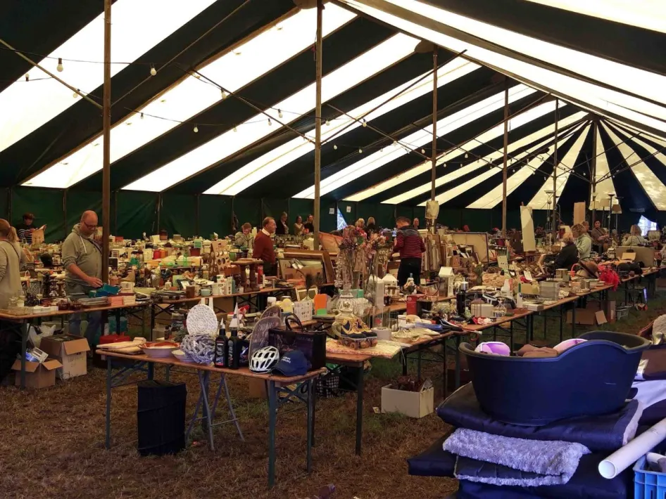 Bezoekers zoeken tussen tweedehands spullen tijdens de overdekte vlooienmarkt in een tent.