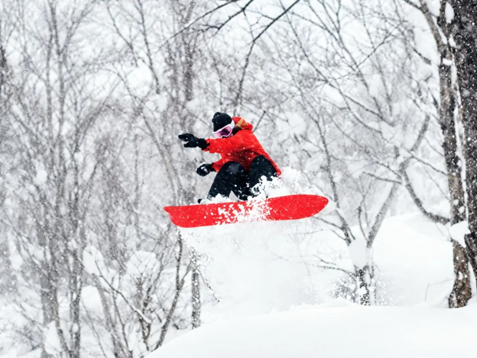Snowboarder in rode jas springt door diepe poedersneeuw in een besneeuwd bos tijdens hevige sneeuwval.