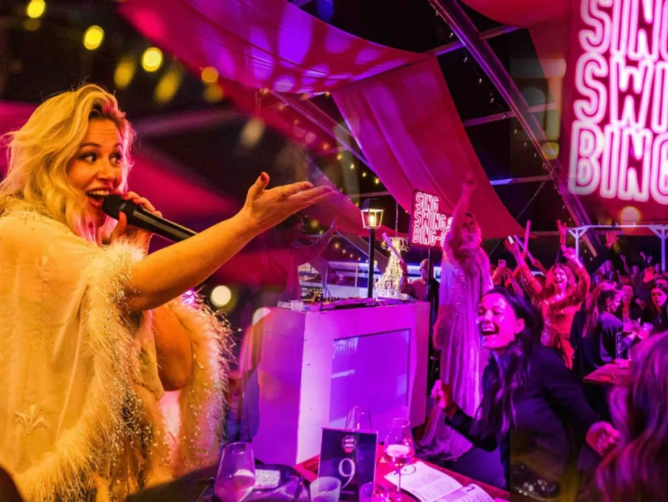 Zangeres met microfoon tijdens Sing, Swing & Bing-o-Long, terwijl het publiek meezingt en feest onder roze en paarse verlichting.