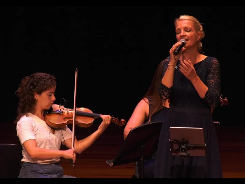 Zangeres Willeke Simons zingt met een microfoon naast een violiste op het podium.
