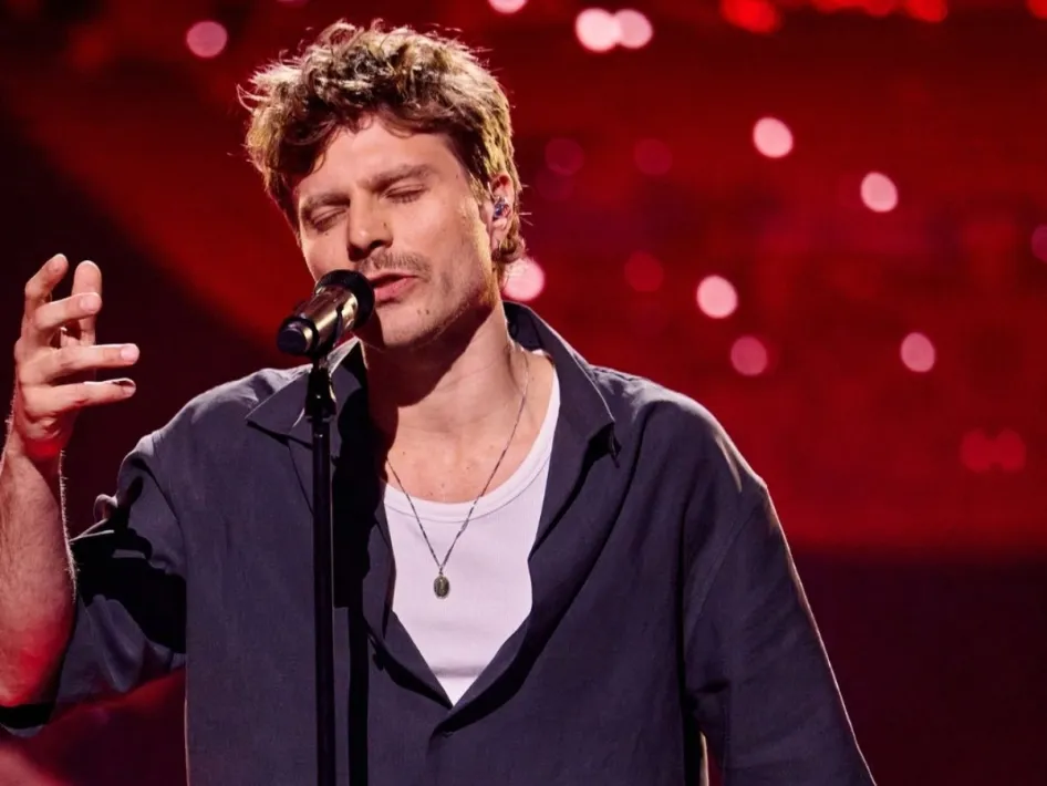 Charl Delemarre zingt met gesloten ogen in een microfoon op een podium met rode verlichting.