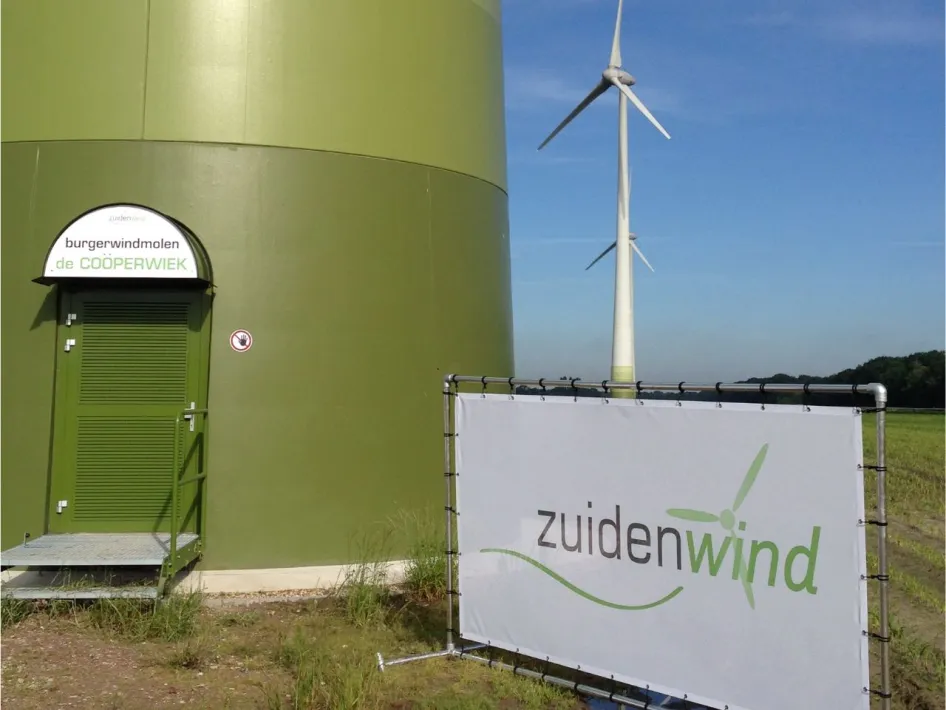 Voet van burgerwindmolen De Coöperwiek met een bord van Zuidenwind en een windturbine op de achtergrond.
