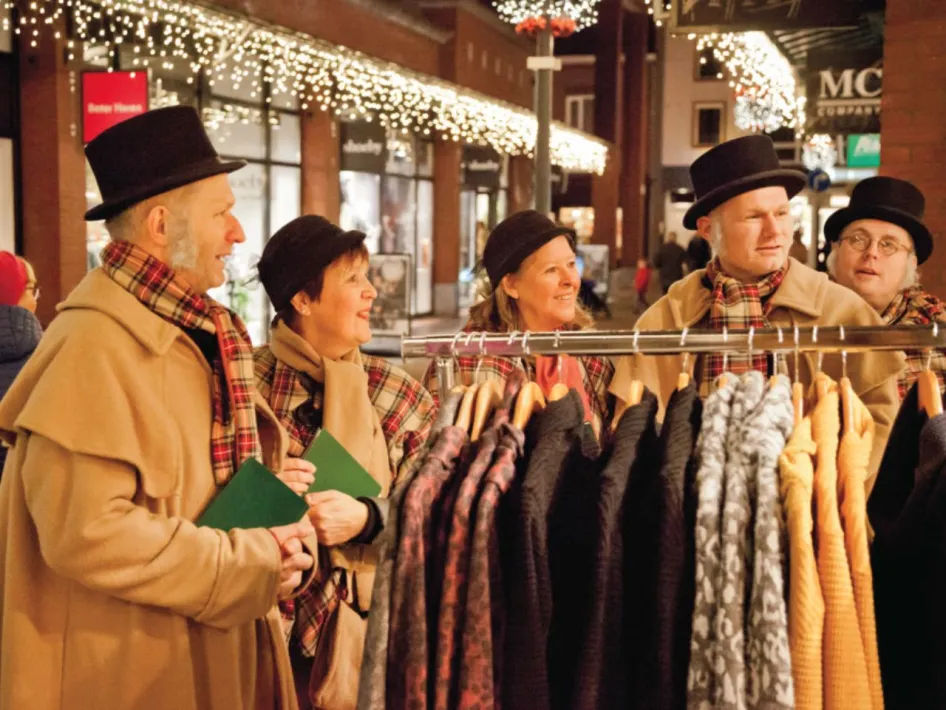 Vijf personen in winterkleding staan bij een kledingrek met jassen tijdens een feestelijke koopavond in een winkelstraat met kerstverlichting.