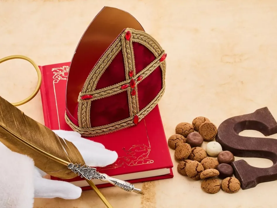 **Alt-tekst:** Sinterklaasitems op tafel met een mijter, rode boek, gouden veer, kruidnoten en een chocoladeletter S.
