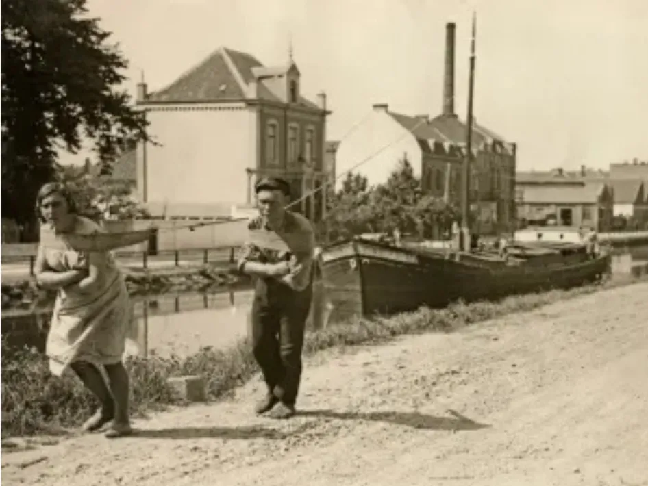 Een jonge vrouw en een man trekken samen een binnenvaartschip voort langs het kanaal — een krachtig beeld van voortvarende vrouwen in de binnenvaart.