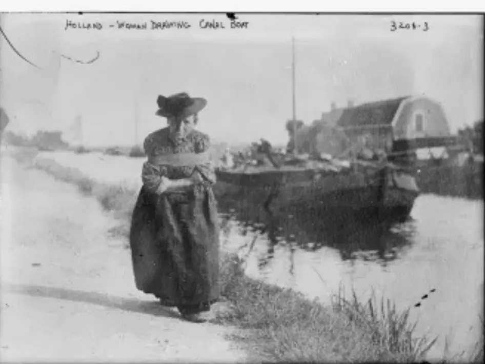 "Vrouw loopt langs een kanaal in Nederland terwijl ze een sleepkabel vasthoudt om een vrachtschuit voort te trekken; op de achtergrond is de boot met lading en een boerderij te zien."