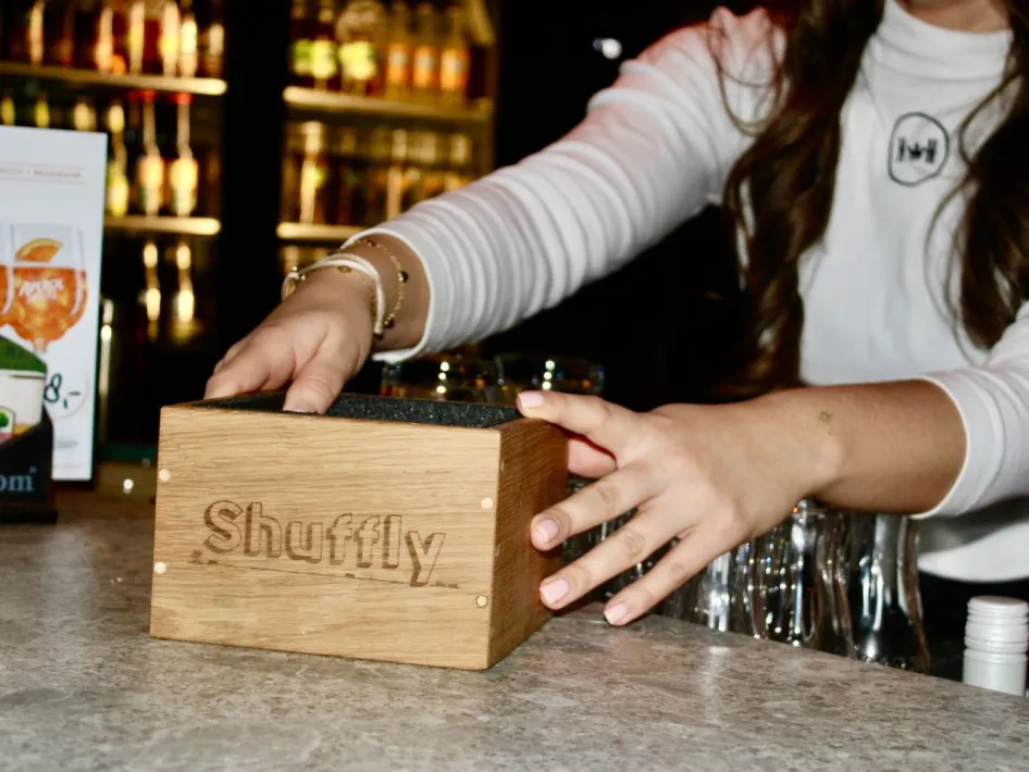 Eine Mitarbeiterin stellt eine Holzbox mit der Aufschrift 'Shuffly' auf die Bar, im Hintergrund ist eine gut bestückte Getränkeregale zu sehen.