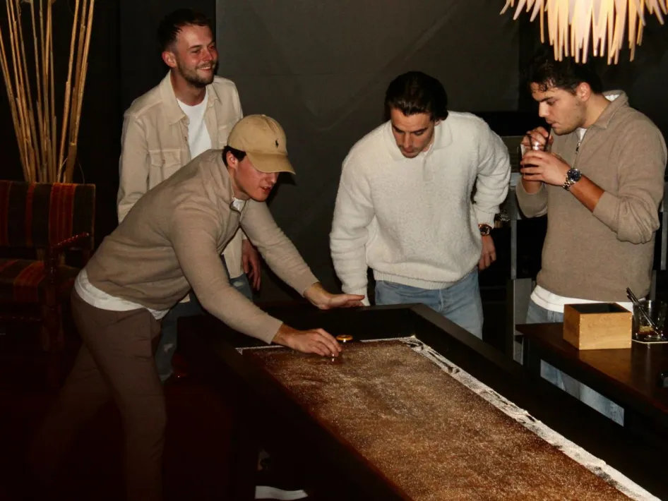 Four young men play American Shuffle in a stylish bar. One player is focused on the game while the others watch and enjoy a drink.