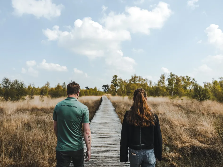 Twee wandelaars op een houten knuppelbrug door het open landschap van De Groote Peel.