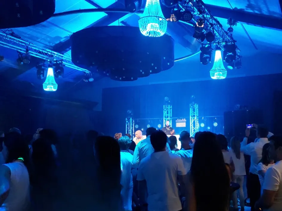 Ein DJ legt auf einer blau beleuchteten Bühne Musik auf, während eine große Gruppe von Gästen in weißer Kleidung unter blau beleuchteten Kronleuchtern tanzt beim Oolderhof.