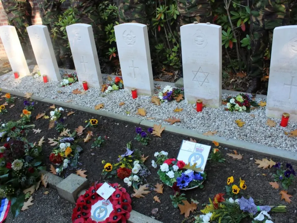 Rij van zes witte oorlogsgraven met herdenkingsstenen, omringd door bloemen, kransen en brandende kaarsen tijdens een herdenkingsplechtigheid.