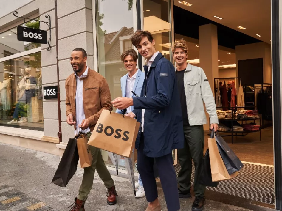 Vier Männer verlassen lachend den BOSS-Store im Designer Outlet Roermond während der Luxury Days, mit mehreren Einkaufstaschen in der Hand.