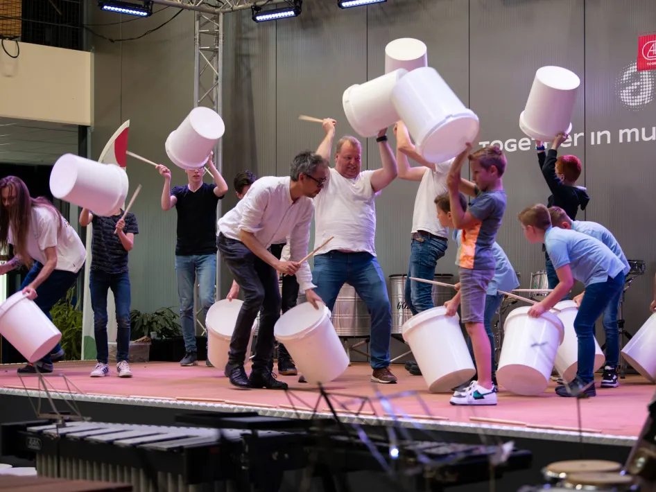 en groep kinderen en volwassenen doet mee aan een interactieve muziekworkshop op het podium, waarbij ze met plastic emmers ritmes maken.