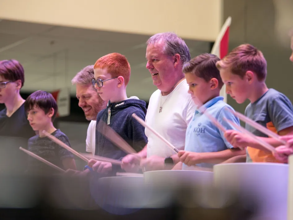 Jongeren en volwassenen staan in een rij tijdens een workshop, geconcentreerd bezig met het bespelen van emmers en slagstokken.