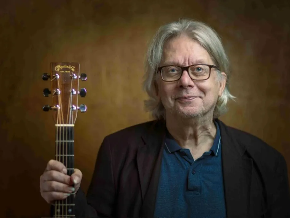 Appeltaartconcert met Ton Engels in het Openluchttheater Thorn. Ton Engels poseert met een akoestische gitaar voor een warme bruine achtergrond.