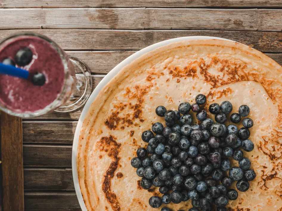Een close-up van een pannenkoek met verse blauwe bessen op een houten tafel, naast een glas blauwe bessensmoothie met een rietje.
