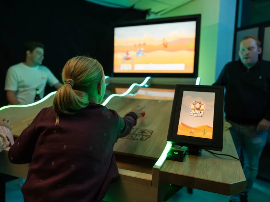 Ein Mädchen spielt ein interaktives Tischspiel mit Animationen auf einem großen Bildschirm, zwei Männer schauen zu.