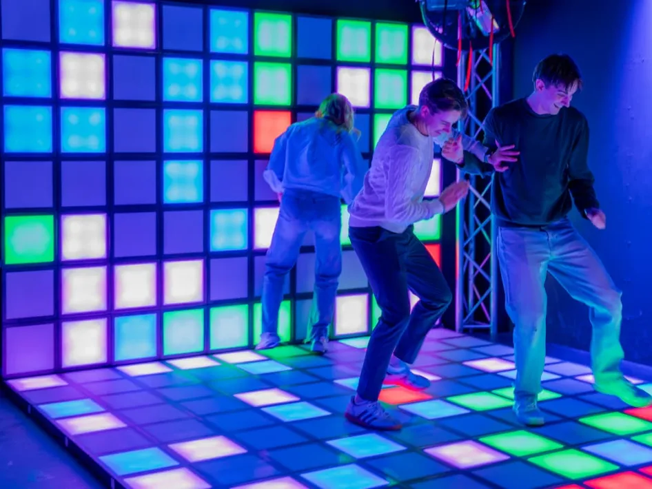 Drie mensen bewegen op een verlichte vloer met gekleurde vlakken in een donkere ruimte bij Poort51 in Weert