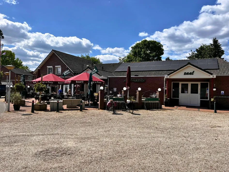 Vorderansicht des Eetcafé de Prairie mit sonniger Terrasse unter roten Sonnenschirmen, Menschen genießen ein Getränk und eine Mahlzeit, neben dem Eingang zum Saal.