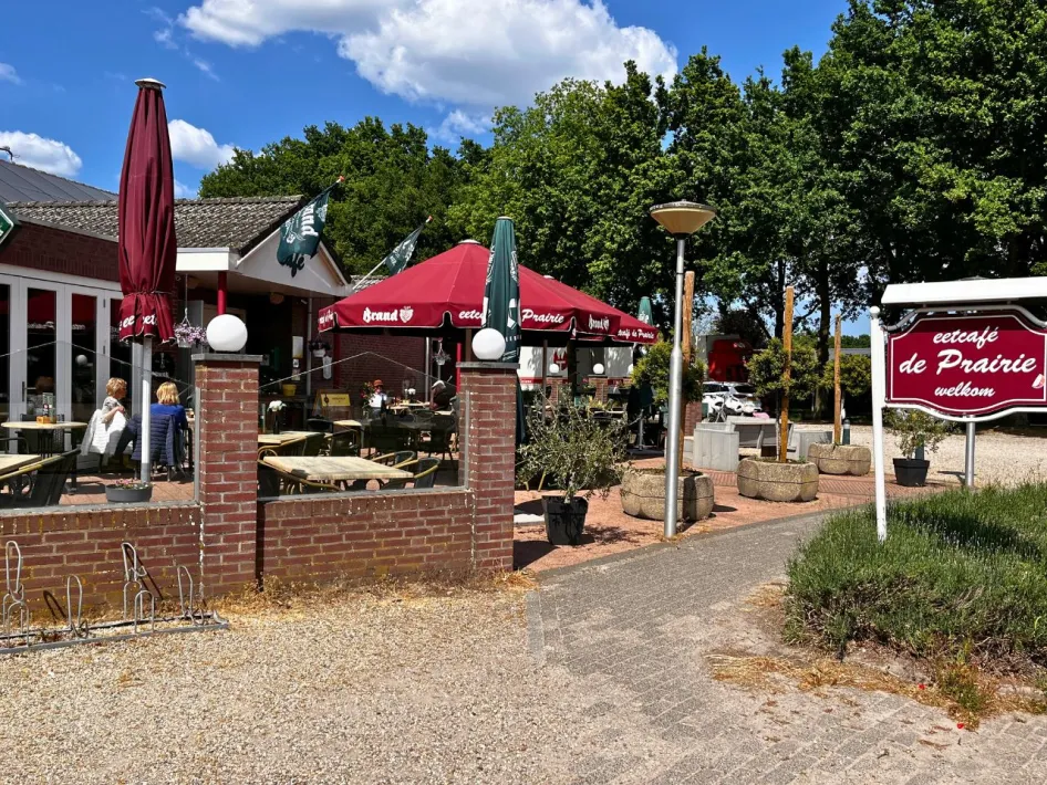 Seitenansicht der Terrasse vom Eetcafé de Prairie mit Gästen unter Sonnenschirmen und einem Willkommensschild am Anfang des Weges.