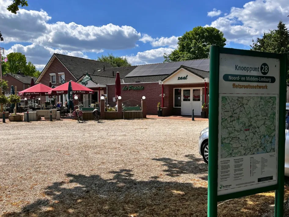 Blick auf das Eetcafé de Prairie unter blauem Himmel mit weißen Wolken, im Vordergrund ein Informationsschild von Fahrradknotenpunkt 28.