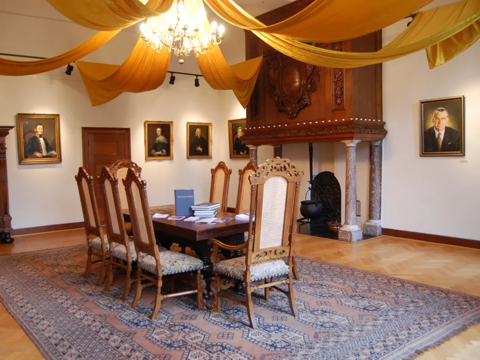 De Regentenkamer in het Prinsenhof in Roermond met antieke houten stoelen en tafel op een oosters tapijt, omringd door klassieke portretten en een rijk gedecoreerde schouw, onder een sierlijk gedrapeerd geel doek en een kroonluchter.