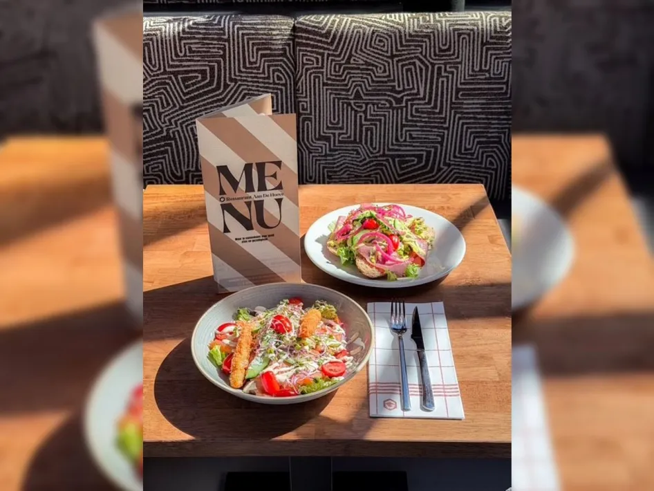 Twee lunchgerechten op tafel bij Restaurant Aan de Hoeve, met salade, brood en een menukaart.