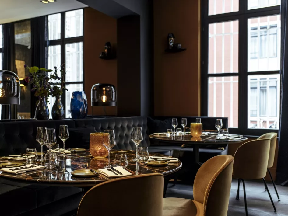 A stylish dining area at Damianz Restaurant, with elegantly set round tables, comfortable chairs and atmospheric lighting. Large windows provide plenty of natural light and a warm ambiance.