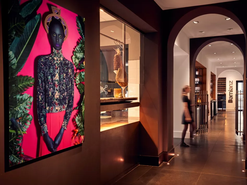 Ein eleganter Korridor im Restaurant Damianz mit einem auffälligen, farbenfrohen Kunstwerk an der Wand, moderner Architektur mit Bögen und einem Blick in die Küche. Der Eingang zum Restaurant ist im Hintergrund zu sehen.