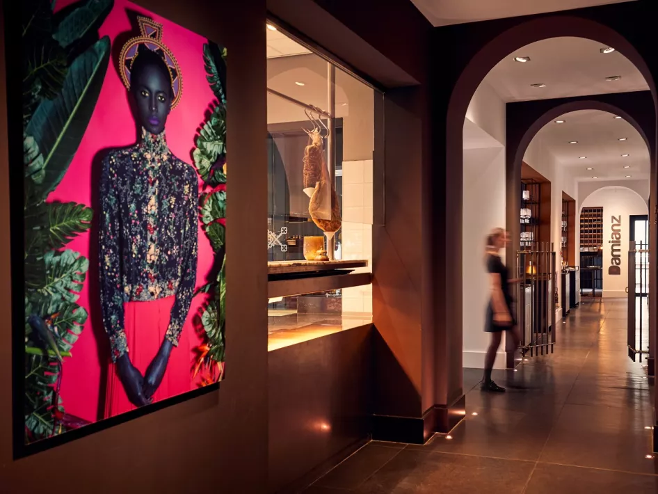 An elegant corridor in Damianz Restaurant, with a striking colorful artwork on the wall, modern architecture with arches, and a view through to the kitchen. The entrance to the restaurant is visible in the background.