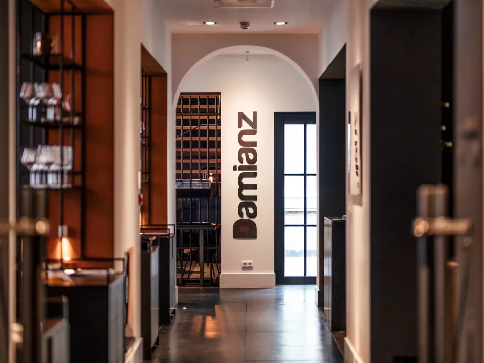 De stijlvolle entree van Restaurant Damianz in Het Arresthuis, met moderne details, warme verlichting en het logo van Damianz prominent op de witte muur aan het einde van de gang.