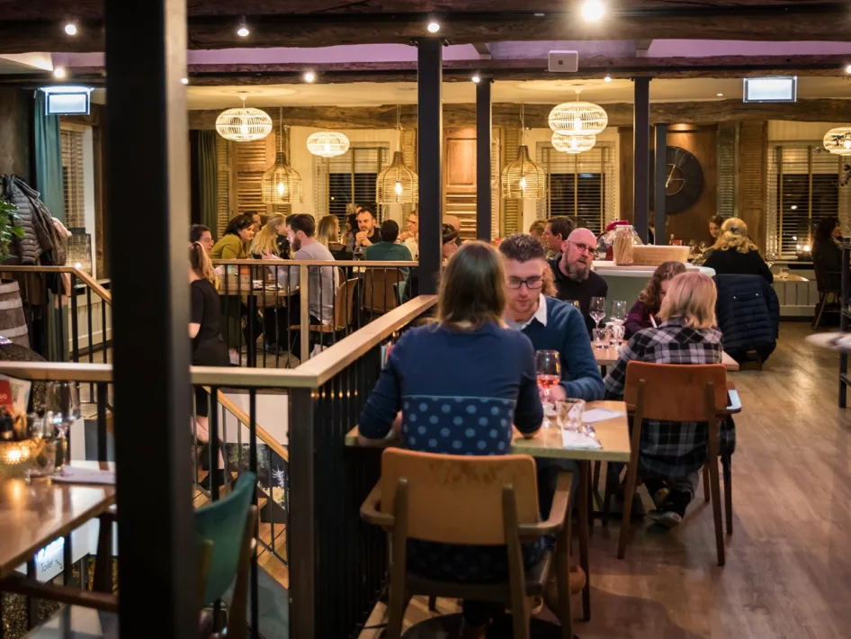 Gasten zitten aan tafels in een sfeervol restaurant met houten balken en warme verlichting.