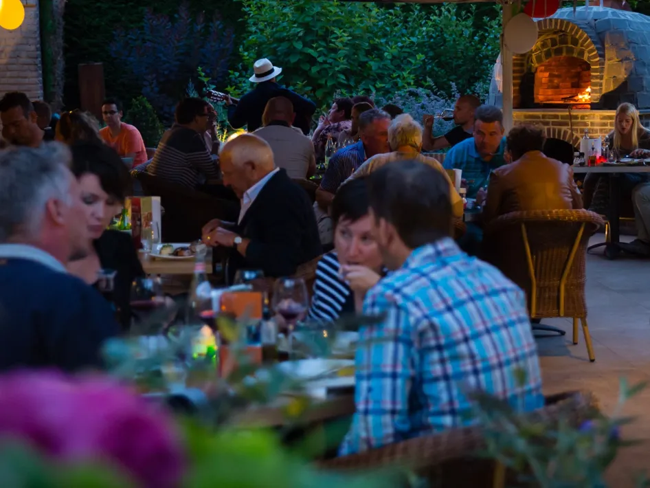 Mensen genieten van eten en drinken op een groen omgeven terras met een buitenhaard.