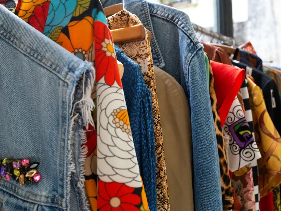 Een kledingrek vol kleurrijke en geprinte kledingstukken, waaronder een spijkerjack met sierstenen en jurken met bloemen- en dierenprints.