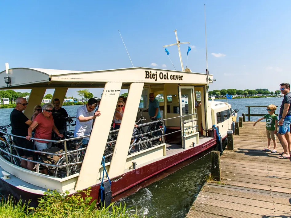 Ferry Biej Ool Euver with cyclists and walkers on board docks at the jetty in Roermond on a sunny day.