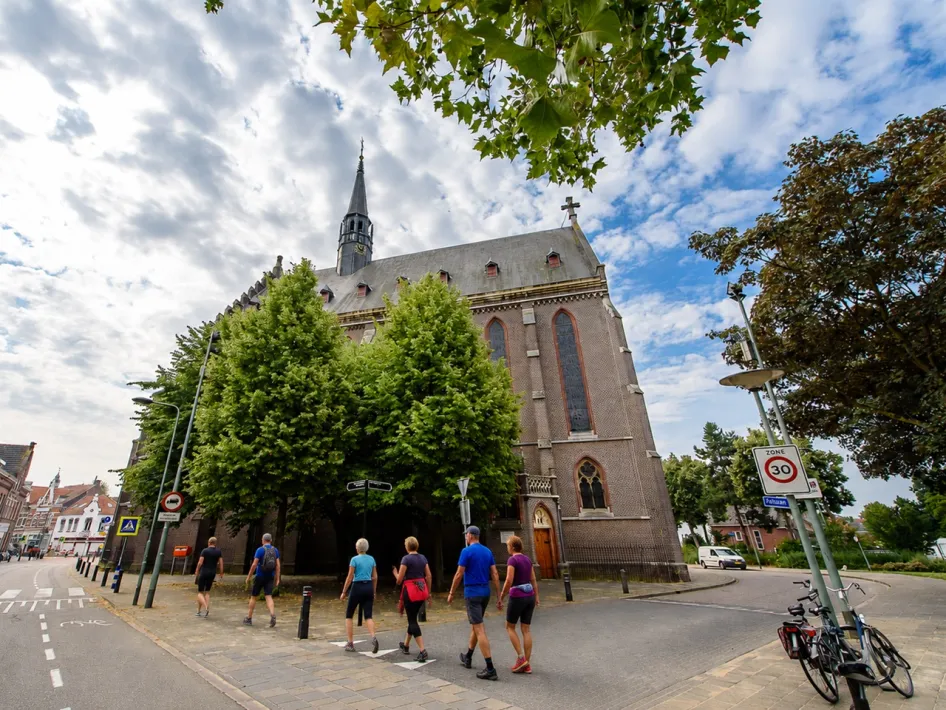 Wandelaars lopen tijdens de wandeltweedaagse Roermond Wandelt langs de Kapel in 't Zand met groene bomen voor de kerk