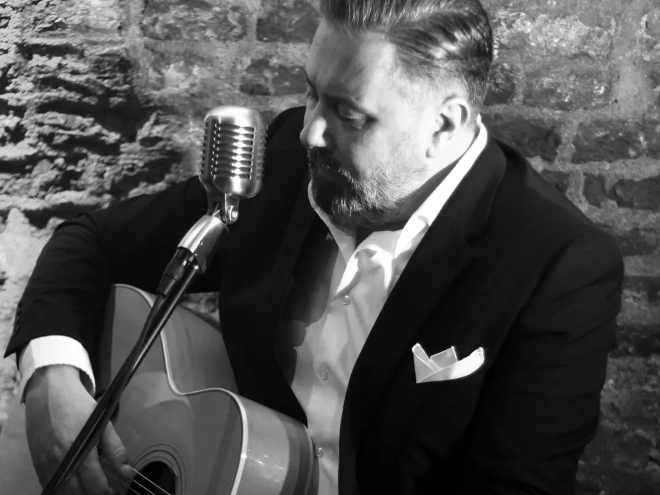 Op een zwart-witte afbeelding speelt Roger Selder zittend gitaar en zingt in een klassieke microfoon