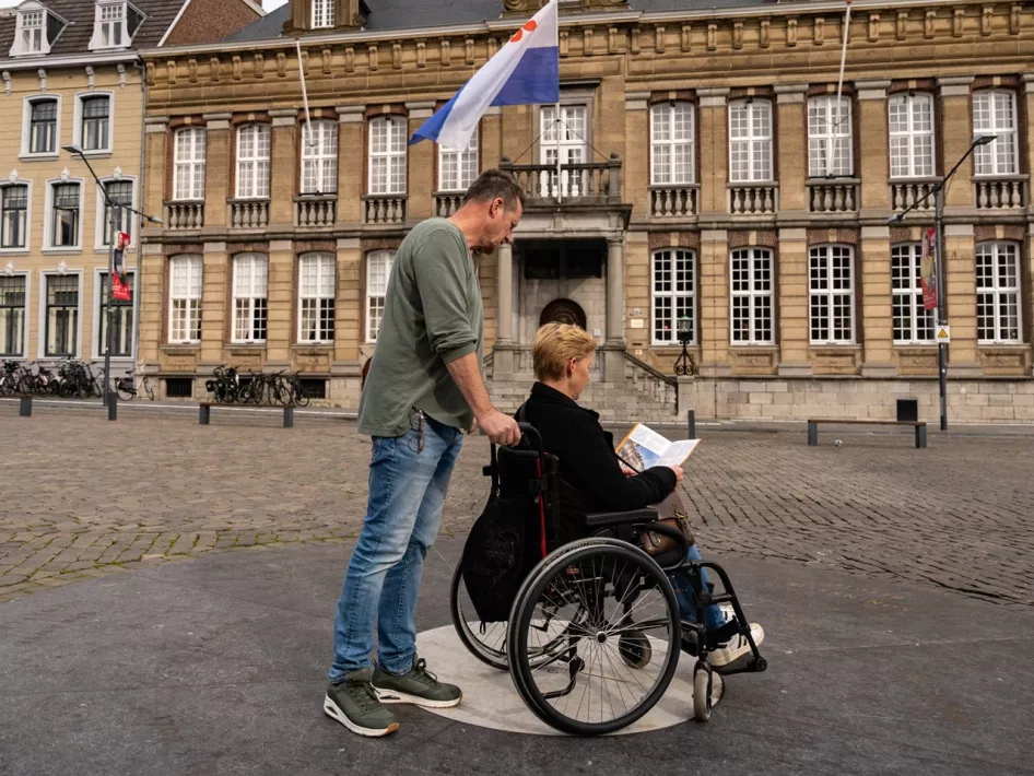 Vrouw in een rolstoel kijkt naar de routekaart van de rolstoelroute - Toegankelijk Roermond terwijl haar man achter haar staat