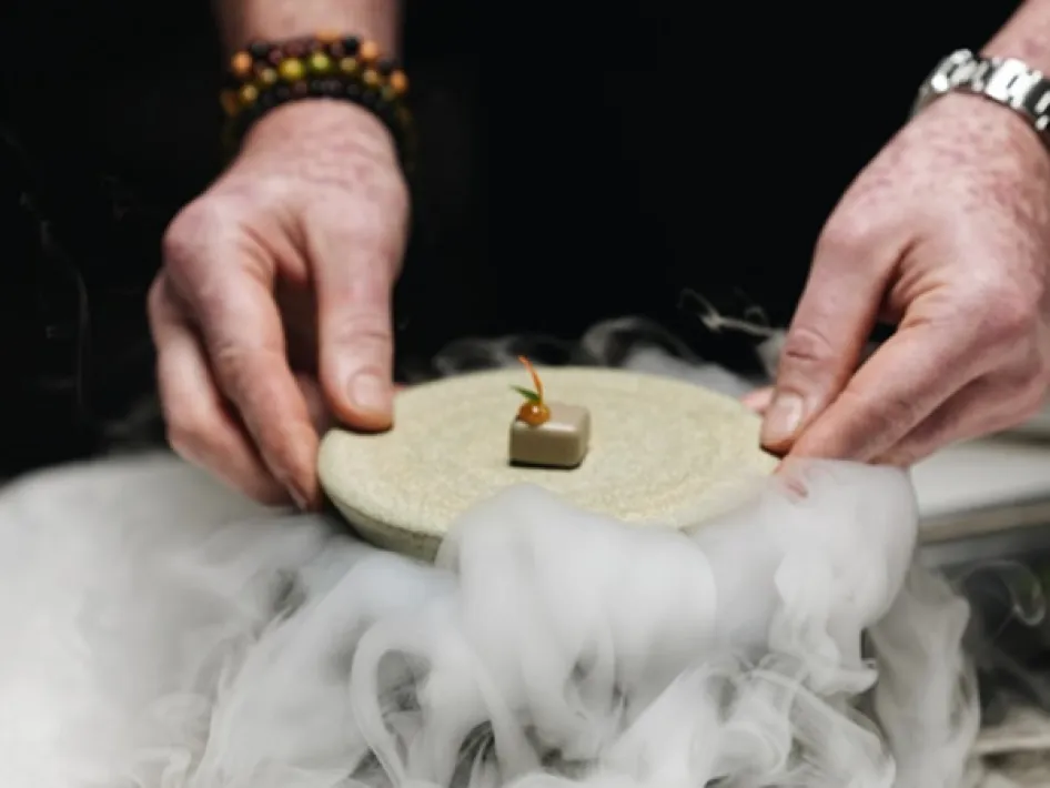 Hands presenting a small dessert on a stone plate above dry ice mist – part of a fine dining experience.