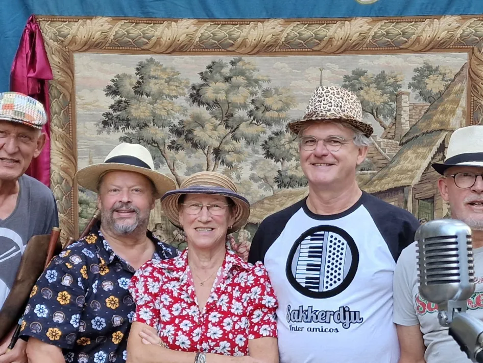 Vijf leden van een muziekgroep poseren vrolijk voor een decoratief wandkleed, met een microfoon op de voorgrond.