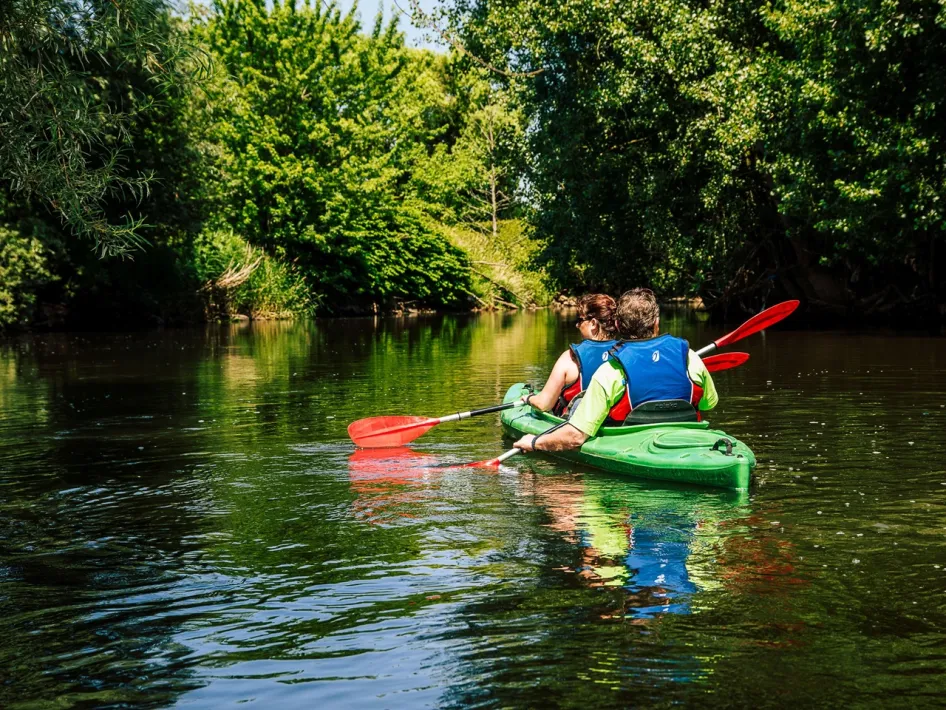Two people canoeing on the Roer at Schurenhof, surrounded by dense greenery and trees along the banks