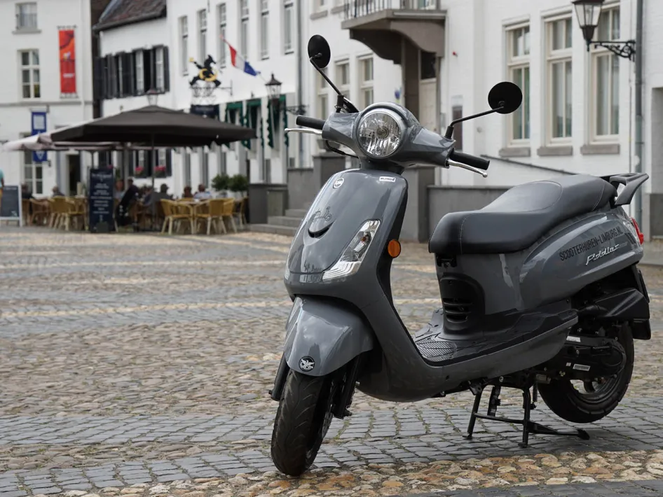 Grijze scooter geparkeerd op een geplaveid plein met witte historische gebouwen en een terras met parasols op de achtergrond.