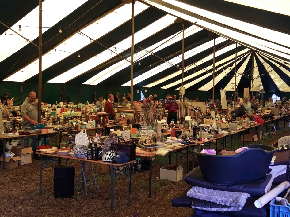 Overzicht van een rommelmarkt in een grote tent met zwart-witte strepen, tafels vol tweedehands spullen en enkele bezoekers die rondkijken.