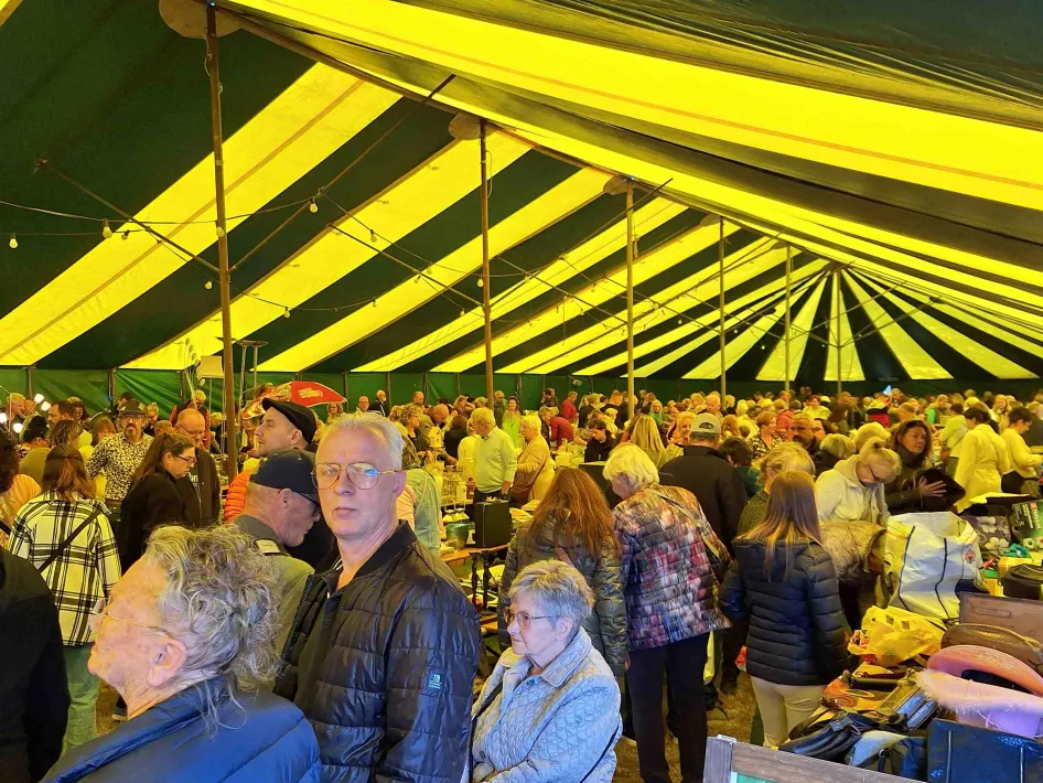 Drukke rommelmarkt in een grote feesttent met geel-groene strepen, waar bezoekers langs de kramen lopen en rondsnuffelen.