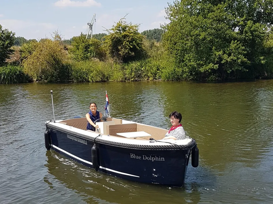 Zwei Personen fahren entspannt mit einer offenen Schaluppe namens Blue Dolphin über einen ruhigen Fluss, umgeben von viel Grün.