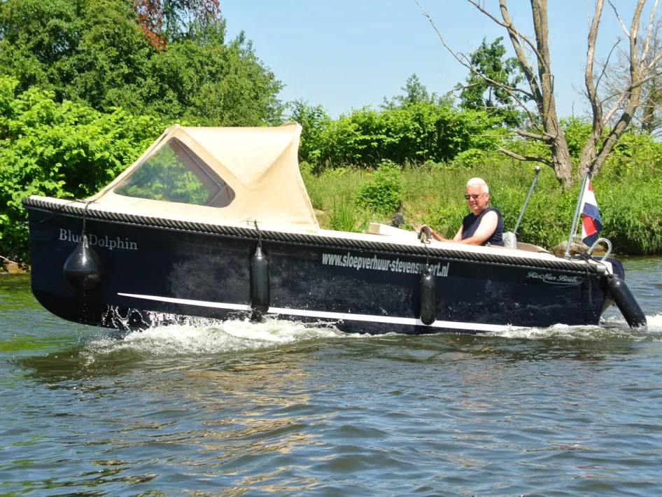 Ein Mann steuert eine Schaluppe von Sloepverhuur Stevensweert mit Sonnendach und niederländischer Flagge, entlang eines grünen Ufers.